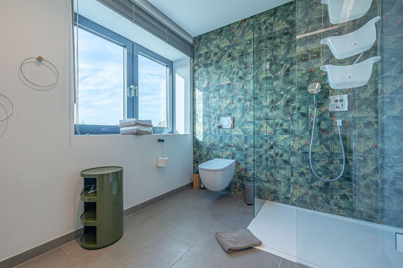 Badezimmer mit Dusche, Toilette und Fenster. Wandfliesen mit tropischem Muster.
