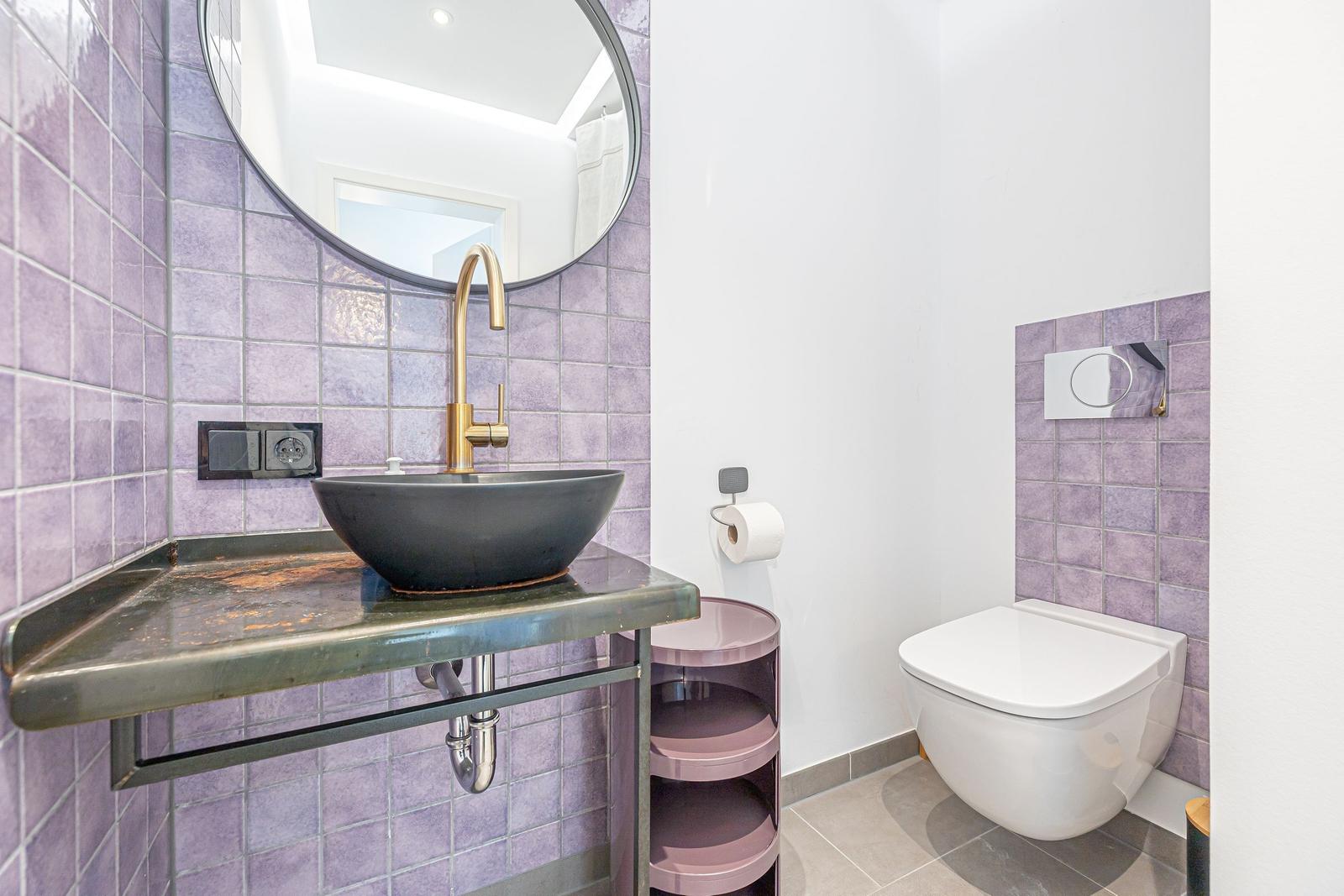 Badezimmer mit Lavendelfliesen, runder Spiegel, schwarzer Waschschale und Wandtoilette.