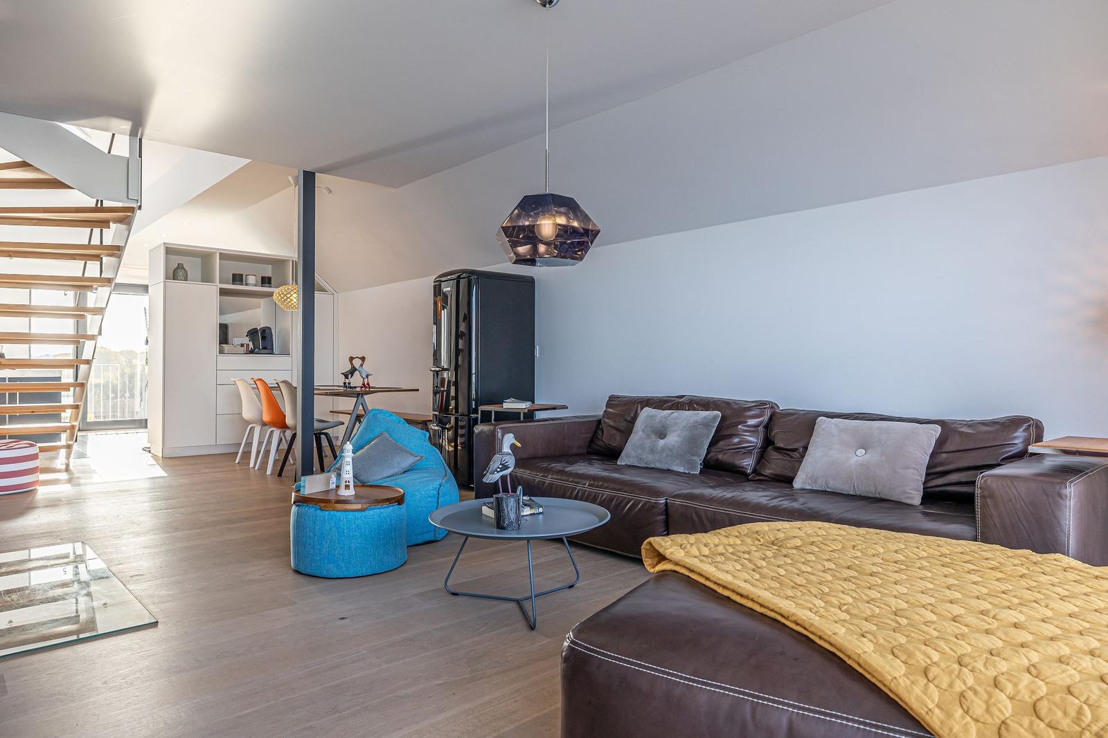 Wohnzimmer mit braunem Sofa, blauem Sessel und Holzboden. Küche und Treppe sind sichtbar.