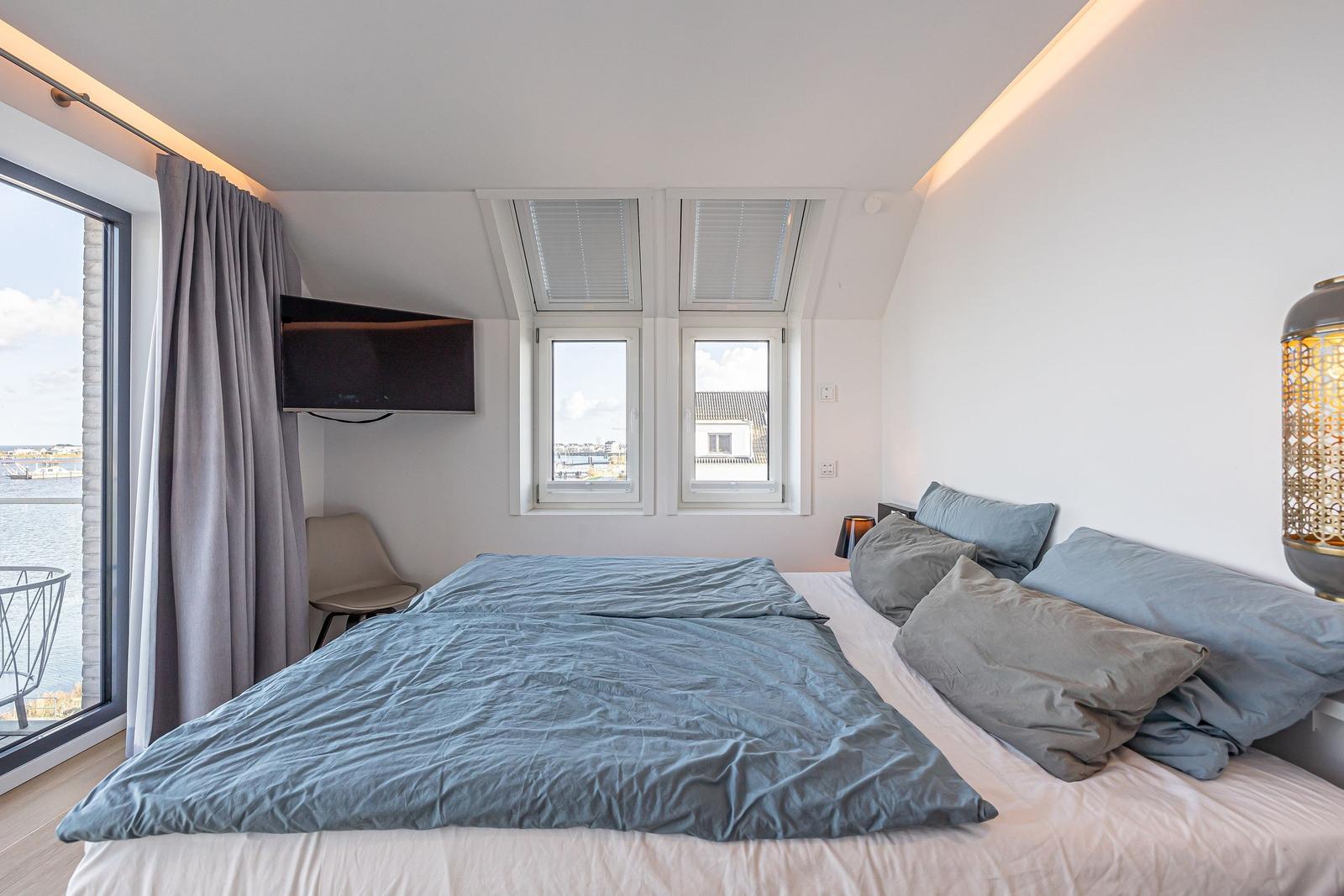 Schlafzimmer mit Bett, Fenstern und TV. Blick auf Wasser durch Glastür.