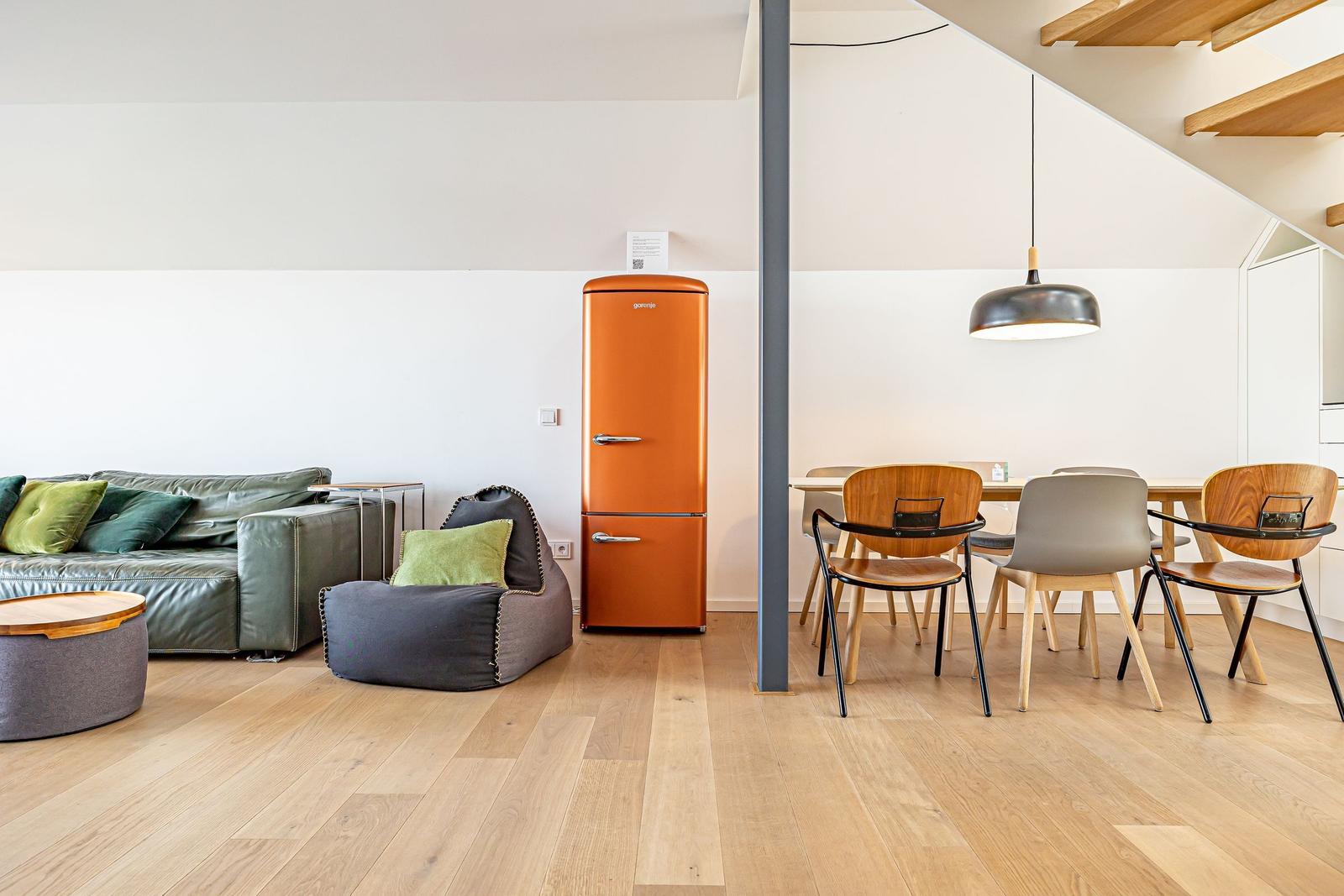 Wohn- und Essbereich mit Sofa, Sitzkissen, orangefarbener Kühlschrank und Holztisch mit Stühlen.