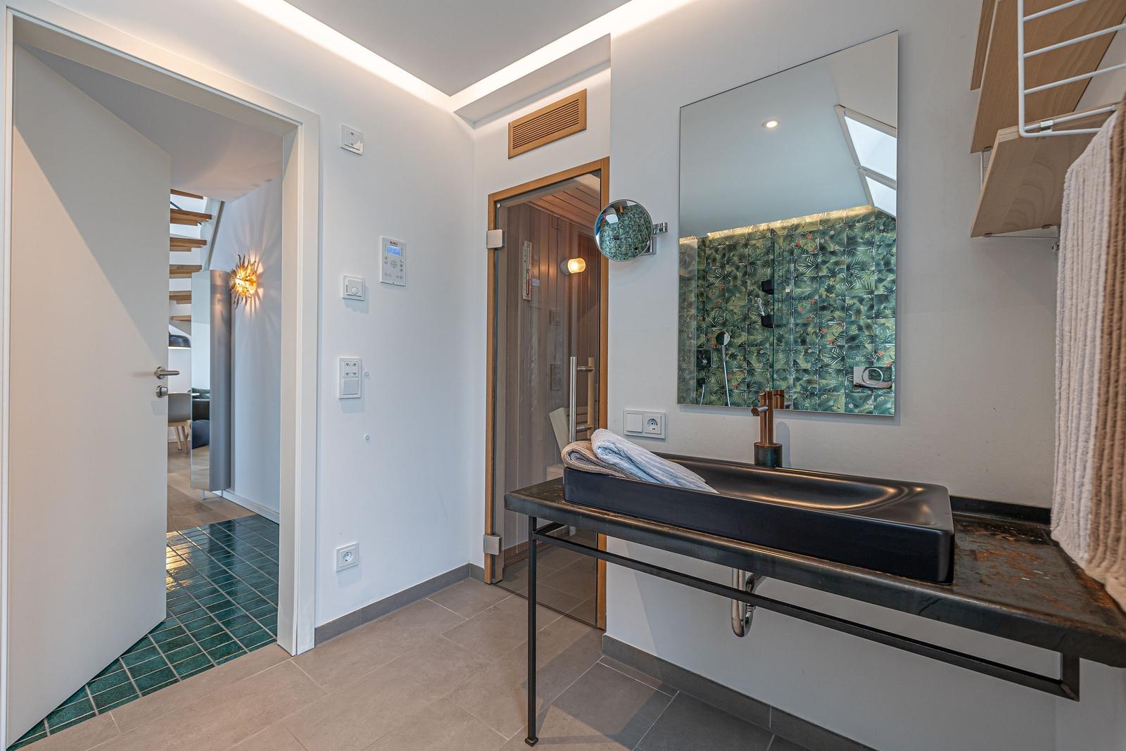 Badezimmer mit Waschtisch, Spiegel und Dusche. Tür führt in Wohnbereich.