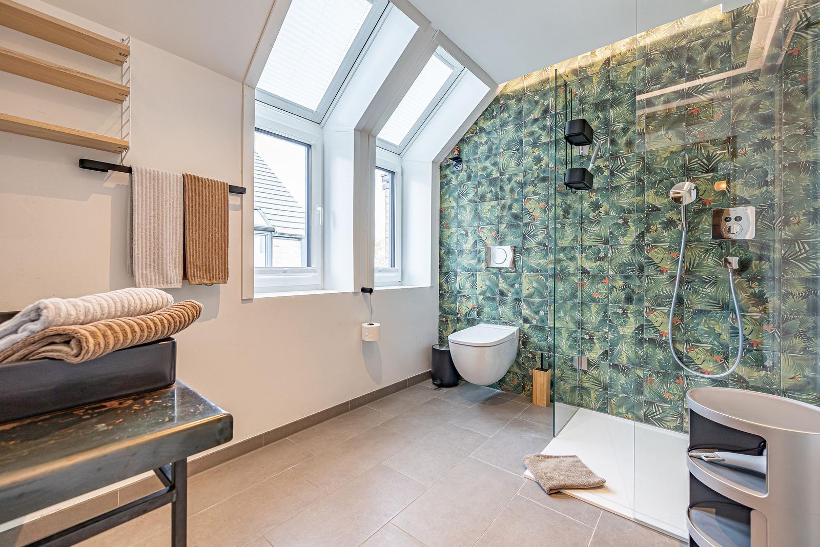 Badezimmer mit Dusche, Toilette und Fenster. Wand mit tropischen Motiven.