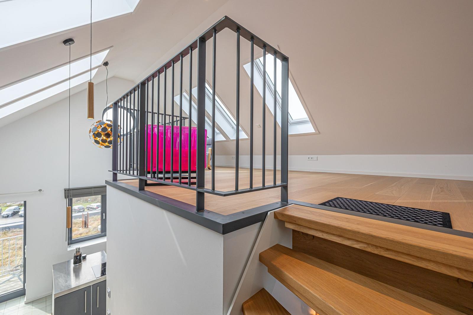 Helle Holztreppe führt zu einer Galerie mit schwarzem Geländer und pinkem Sitzmöbel.