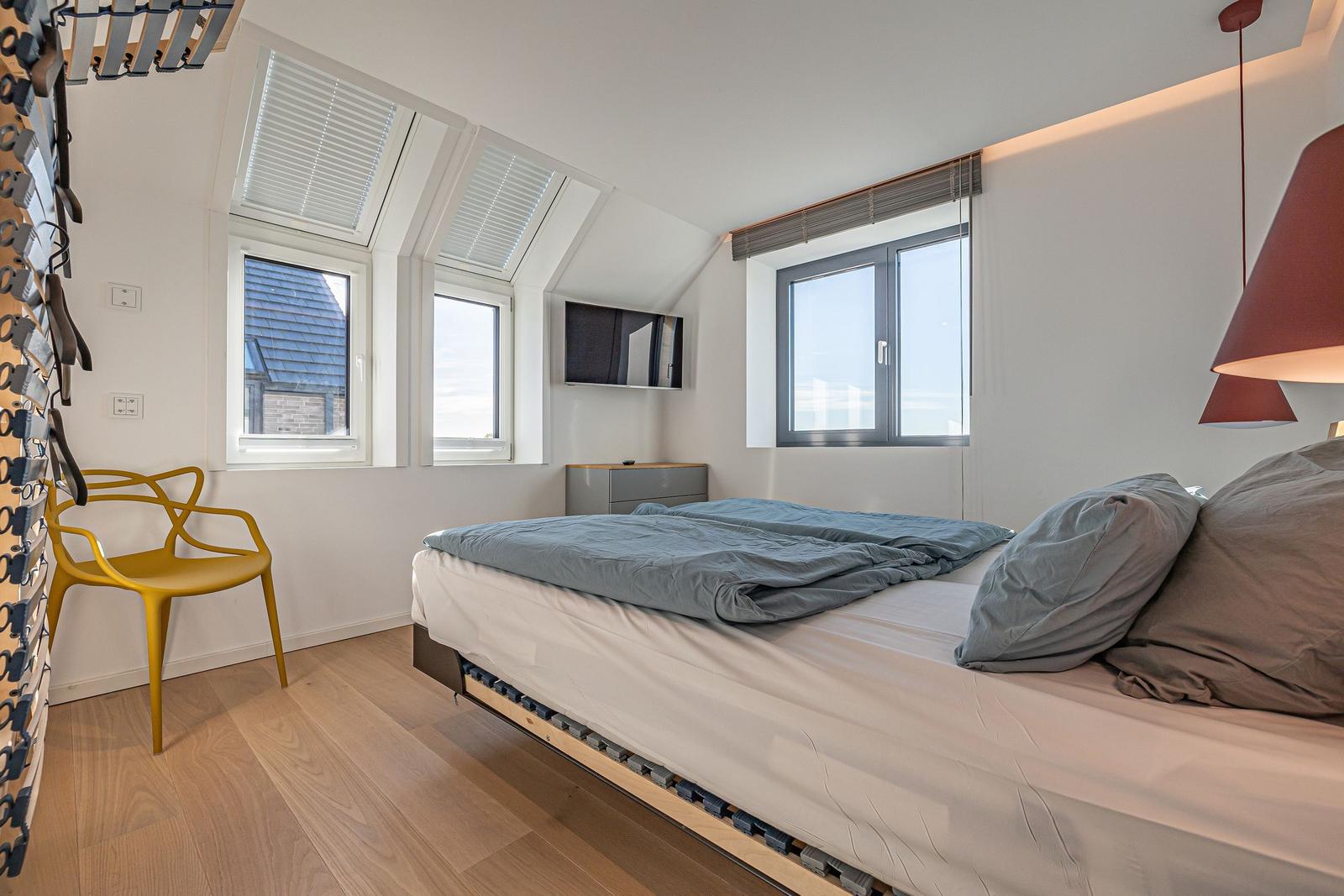Schlafzimmer mit Bett, Fenstern und gelber Stuhl.