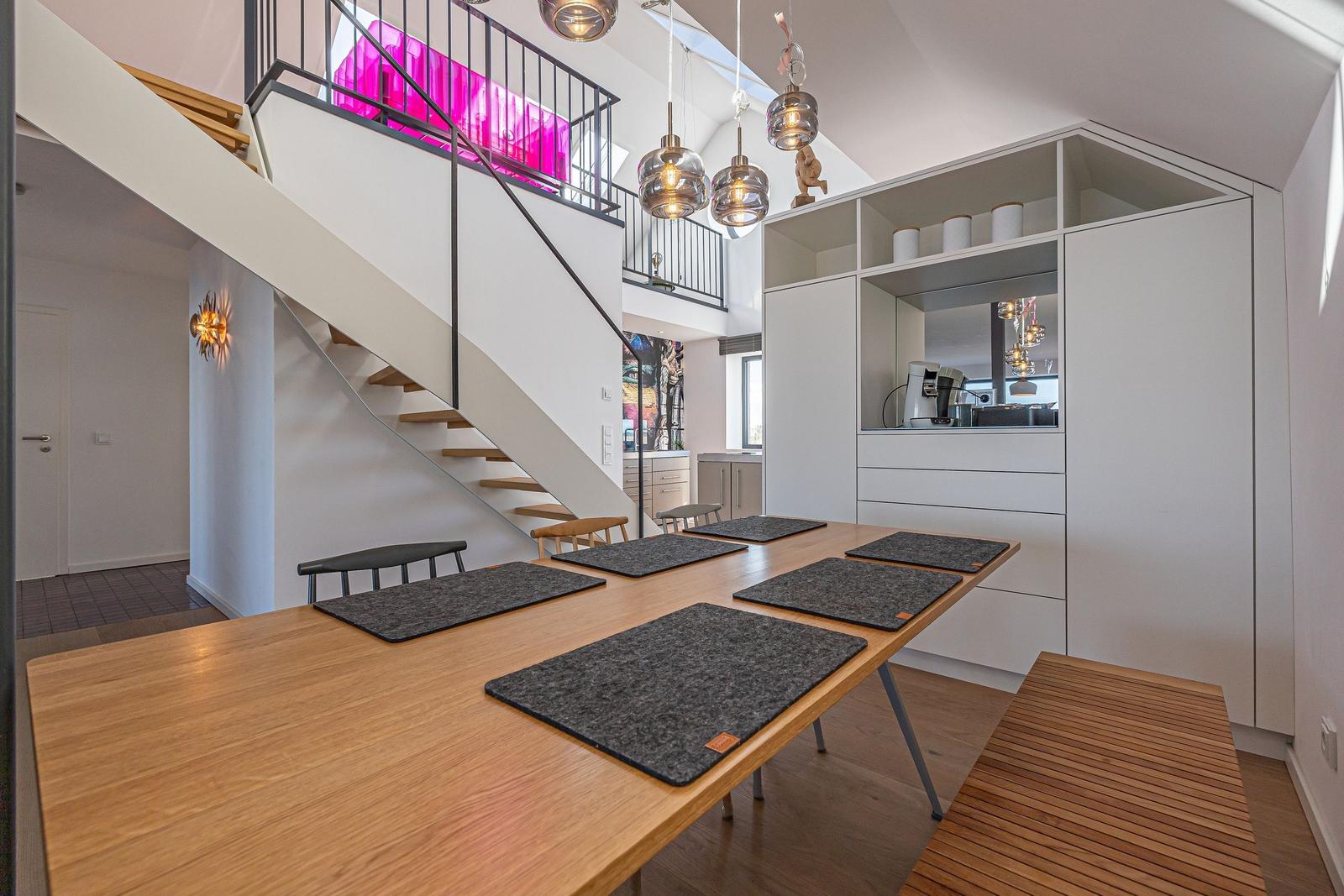 Essbereich mit Holztisch, Stühlen und Platten. Hintergrund: Treppe und Küchenzeile.