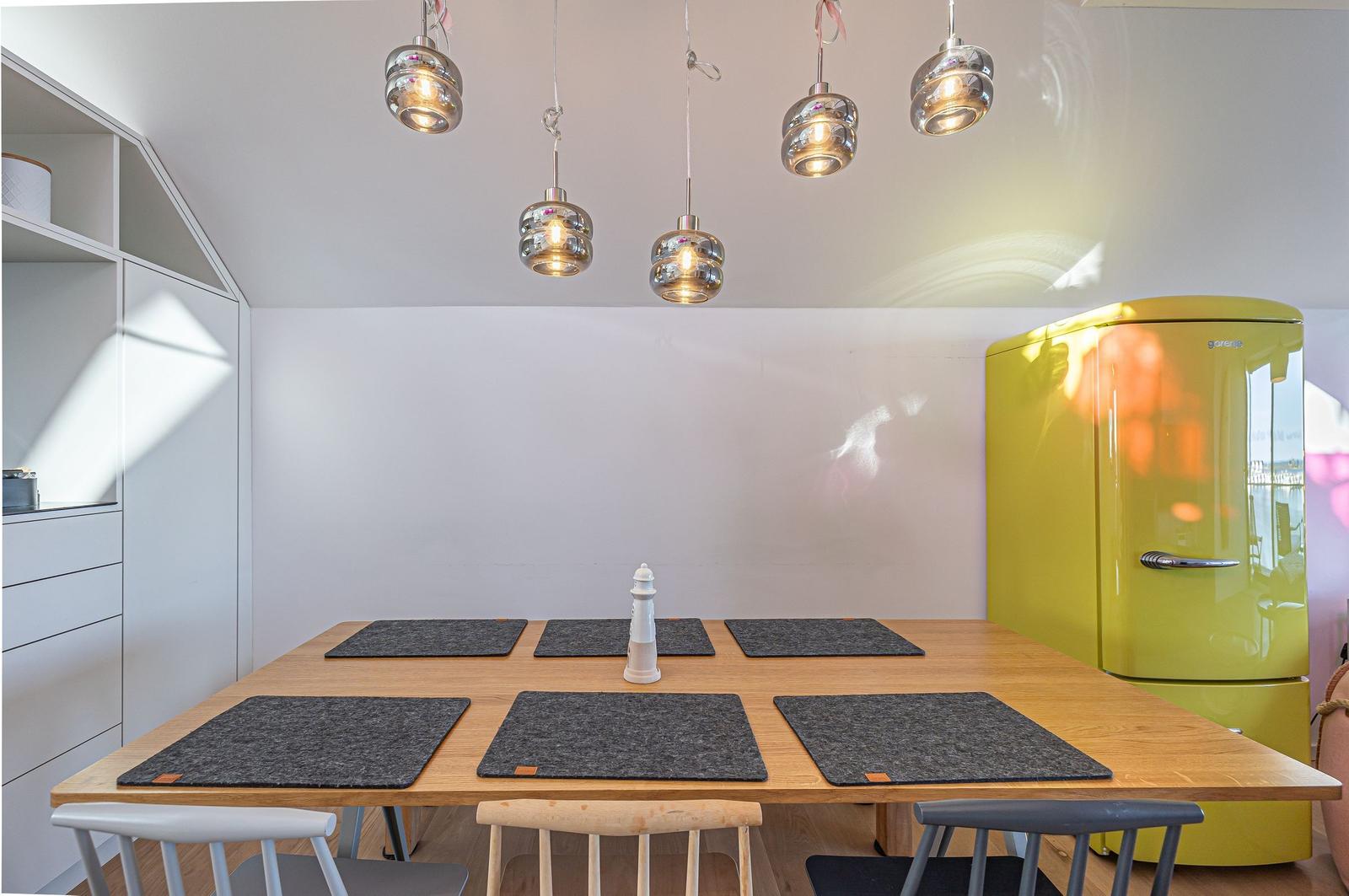Essbereich mit Holztisch, Stühlen und gelbem Kühlschrank. Deckenleuchten und Platten.