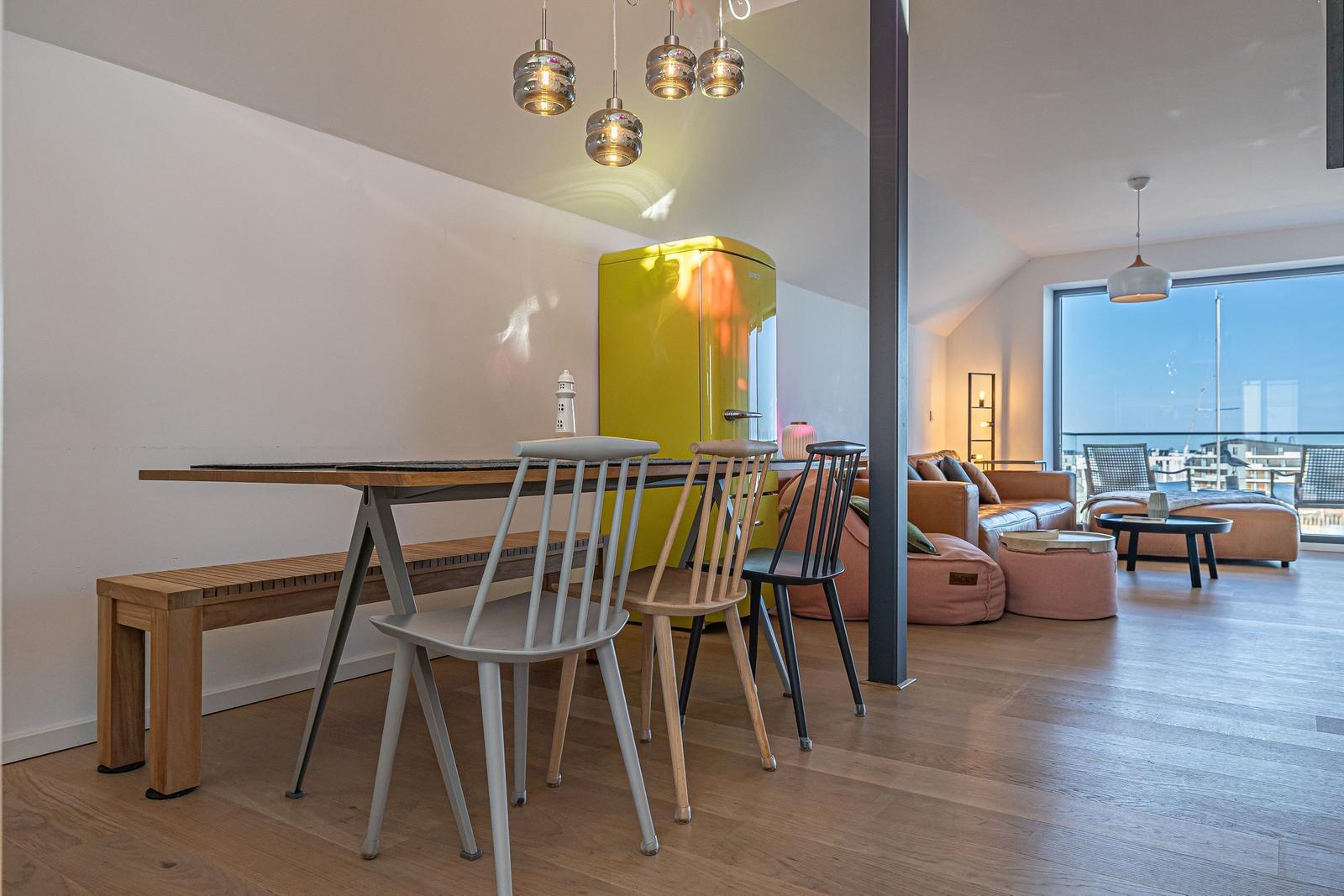 Essbereich mit Tisch, Bänken und Stühlen. Gelber Kühlschrank und Wohnbereich mit Sofa und Blick aufs Meer.