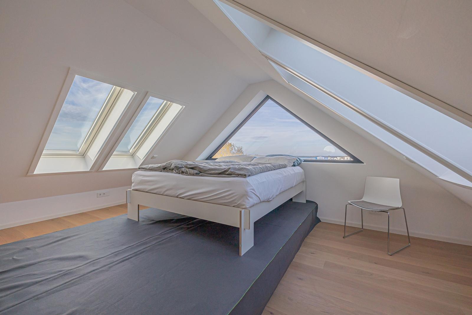 Schlafzimmer mit Dachfenstern und weißem Bett. Holzboden und Stuhl sind sichtbar.