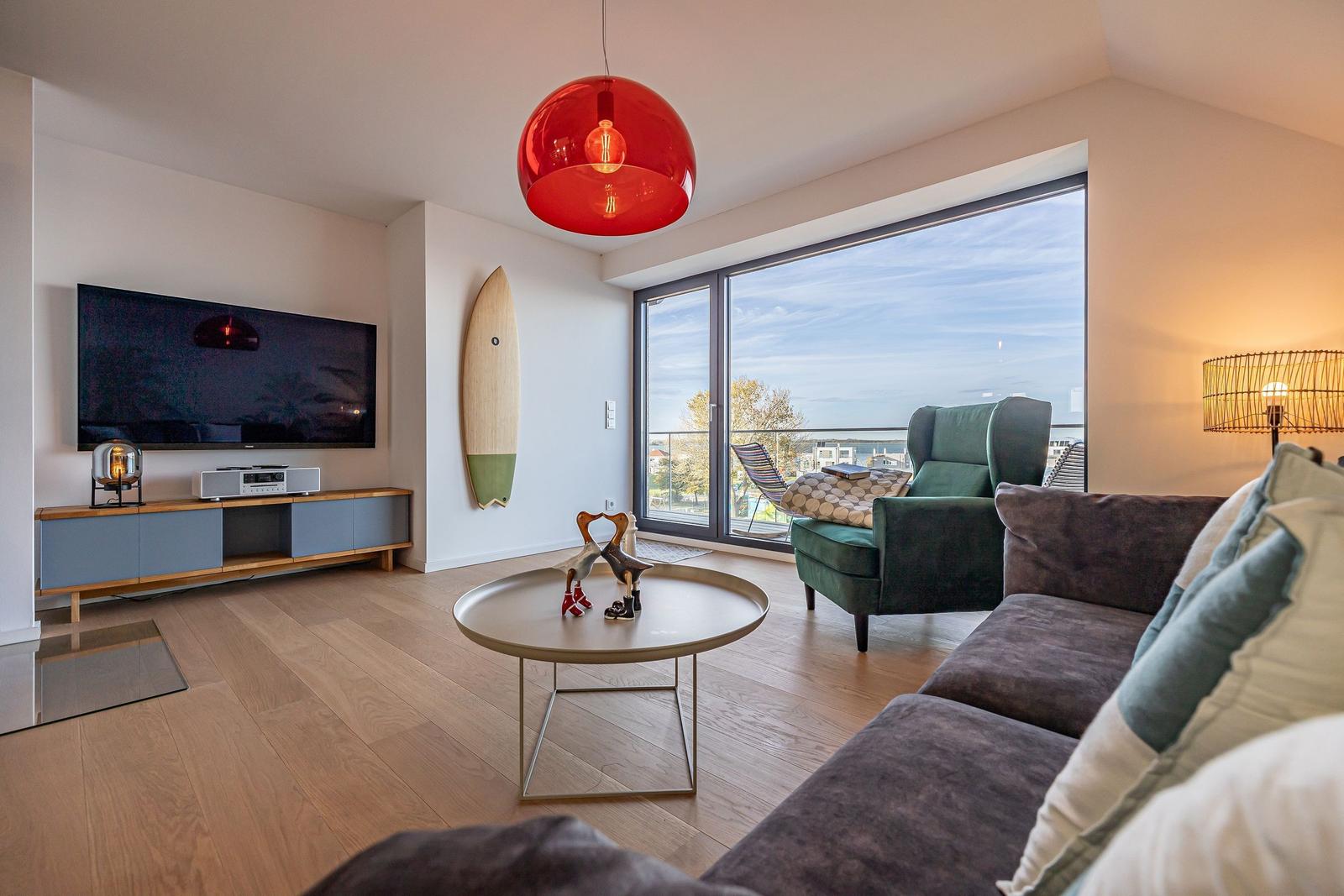Wohnzimmer mit Sofa, Sessel und TV. Große Fensterfront mit Aussicht. Roter Hängelampe und Holzboden.