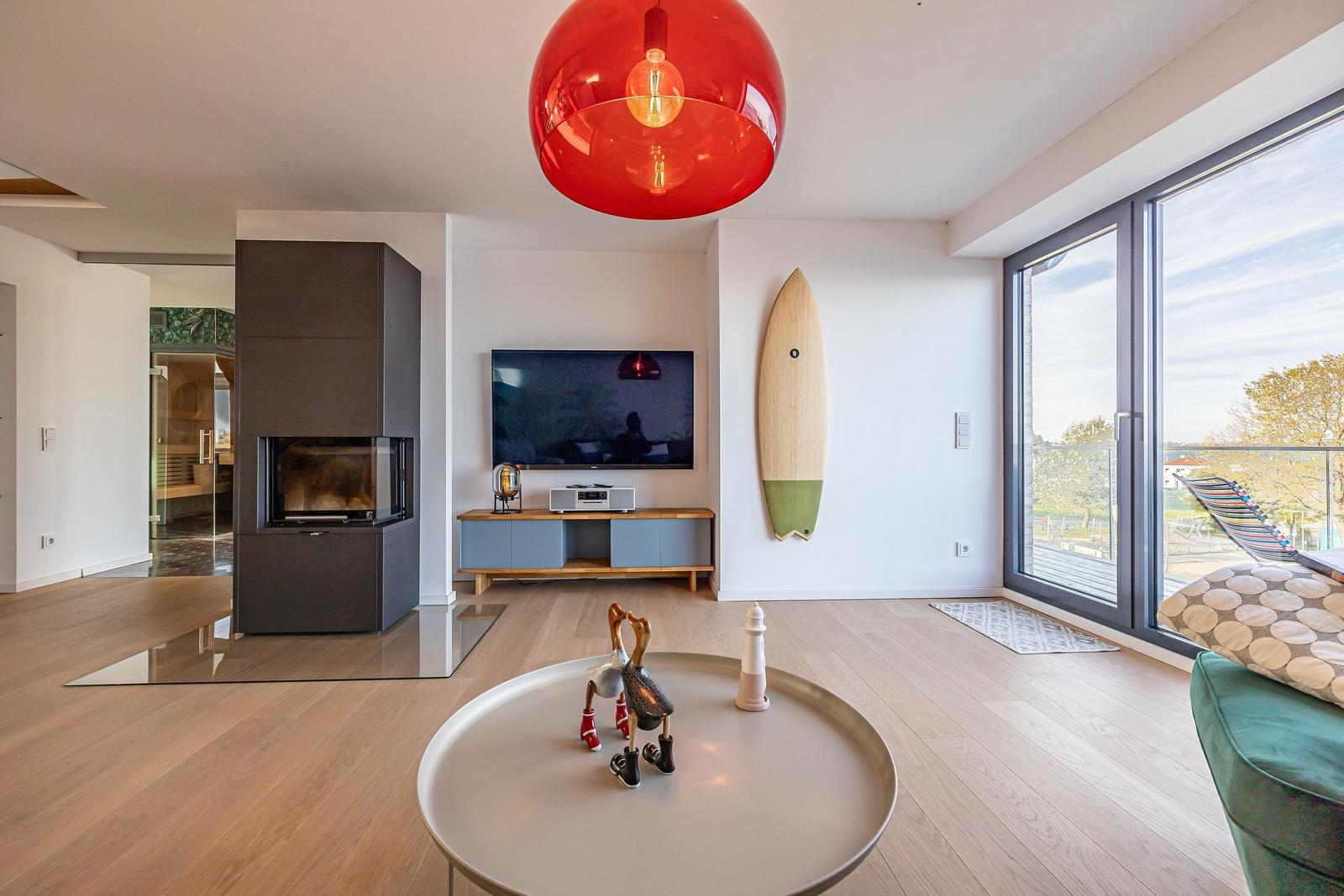 Wohnzimmer mit Kamin, TV, Surfbrett und großem Fenster. Roter Hängelampe und runder Tisch.