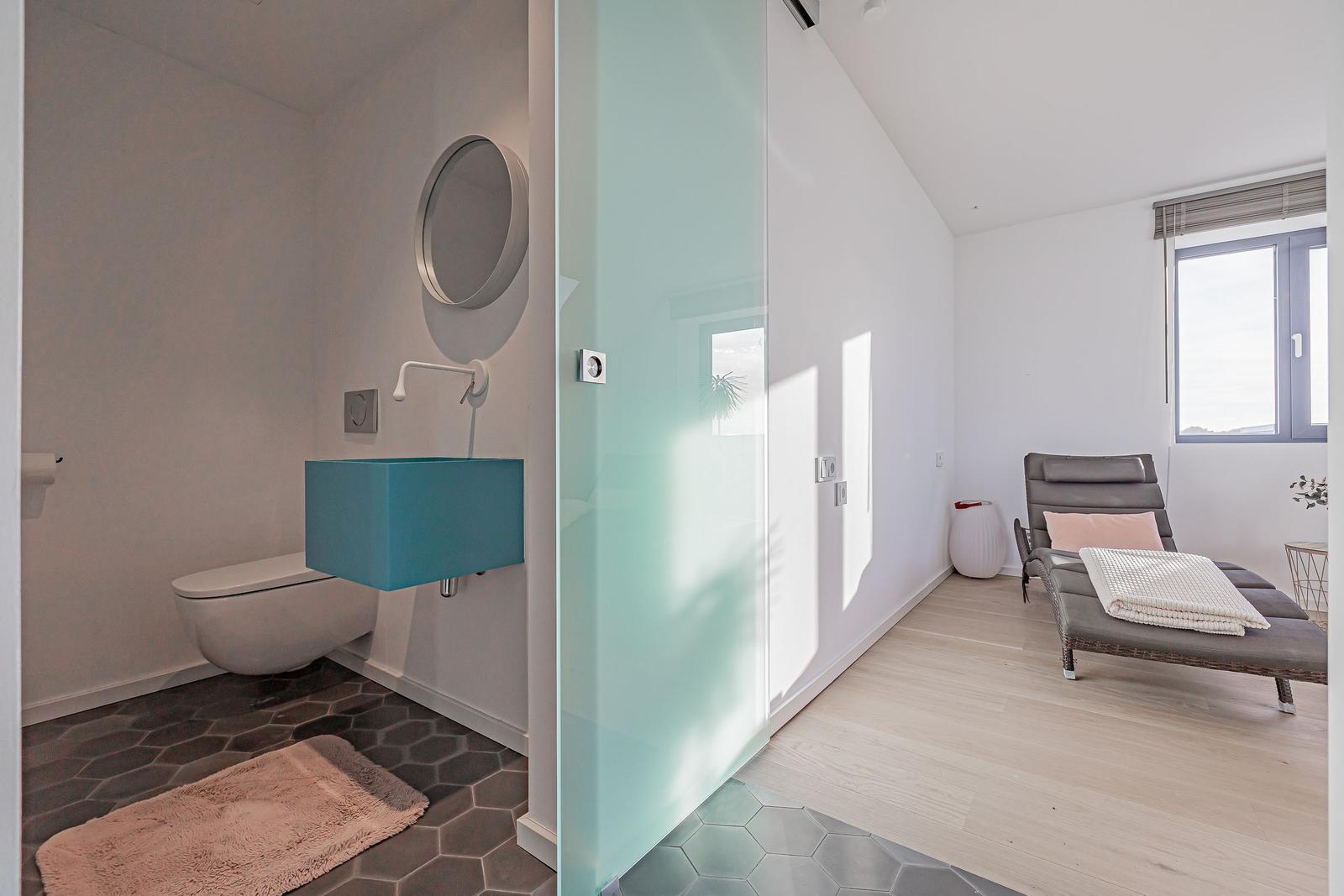 Bad mit Toilette, Waschbecken und Dusche. Nebenraum mit Liege und Fenster.