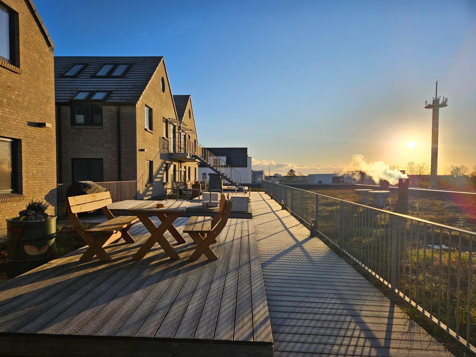 Holzterrasse mit Tisch und Stühlen. Blick auf Gebäude und Sonnenuntergang.