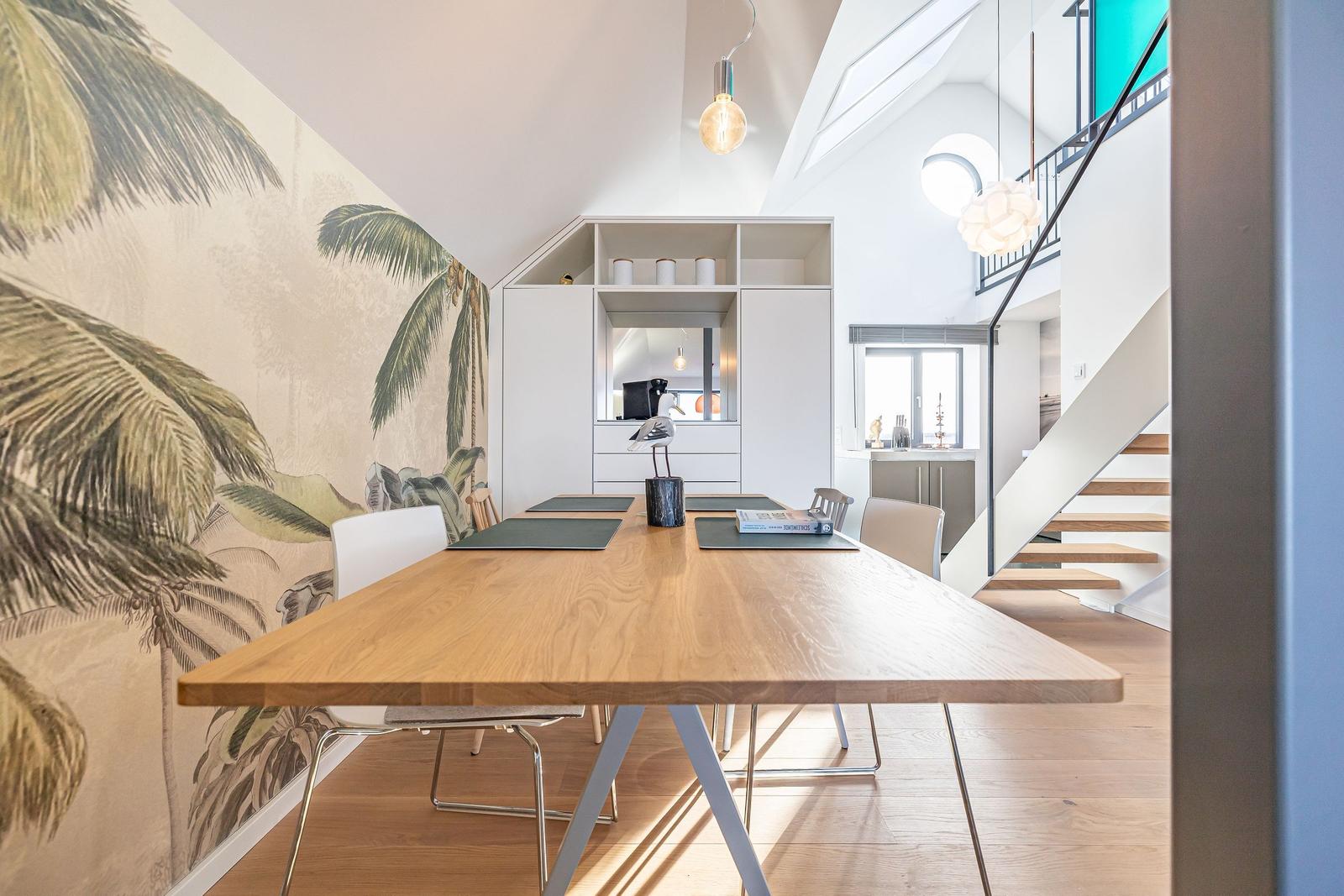 Esszimmer mit Holztisch, Stühlen und Palmenwand. Hintergrund: Küche und Treppe.