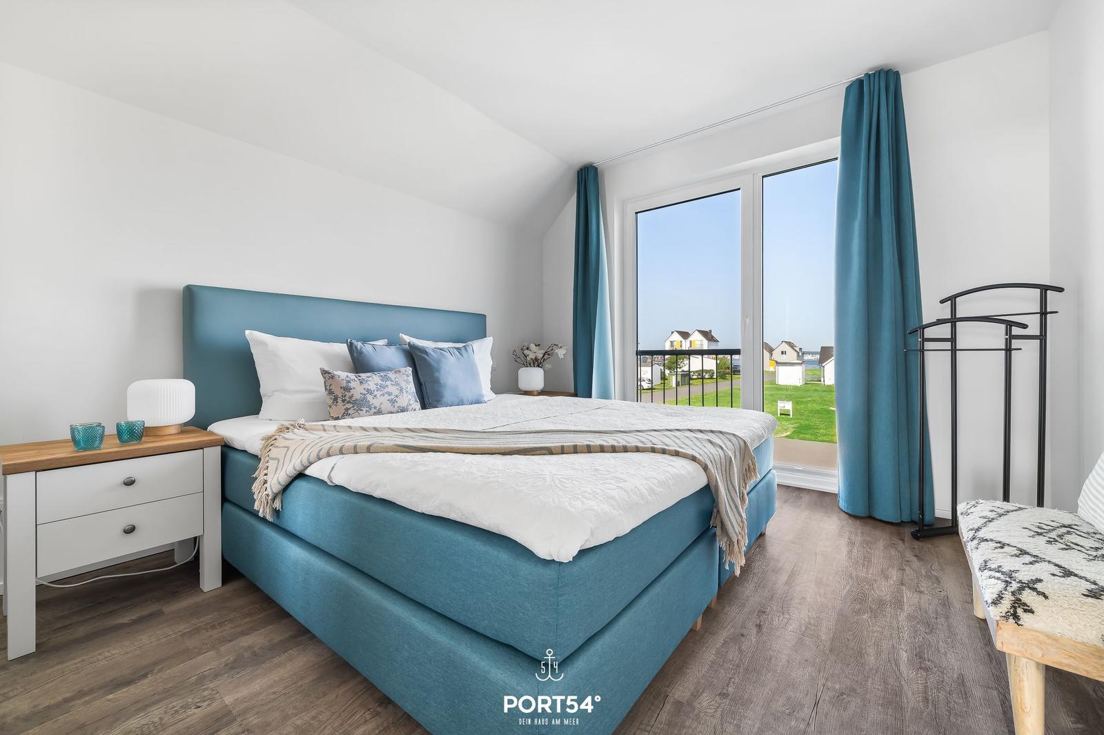 Schlafzimmer mit blauem Bett, großem Fenster und Holzboden.
