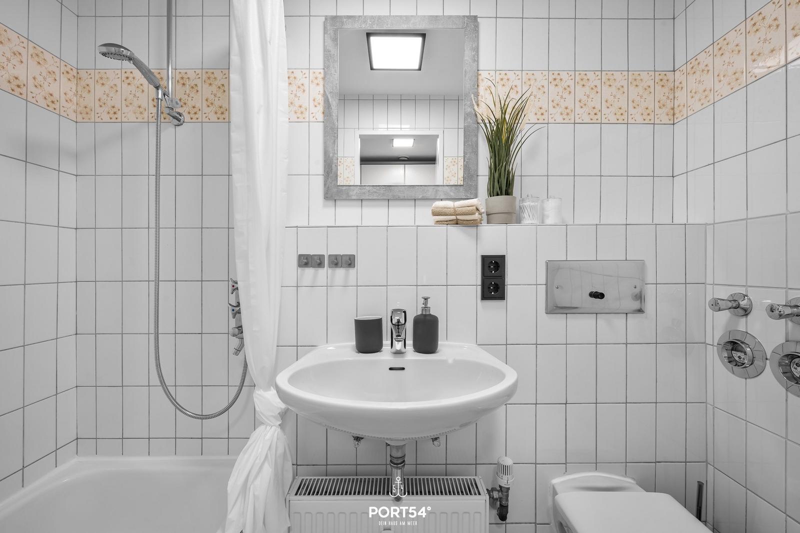 Badezimmer mit Dusche, Waschbecken und Toilette. Weiße Fliesen mit floraler Bordüre.