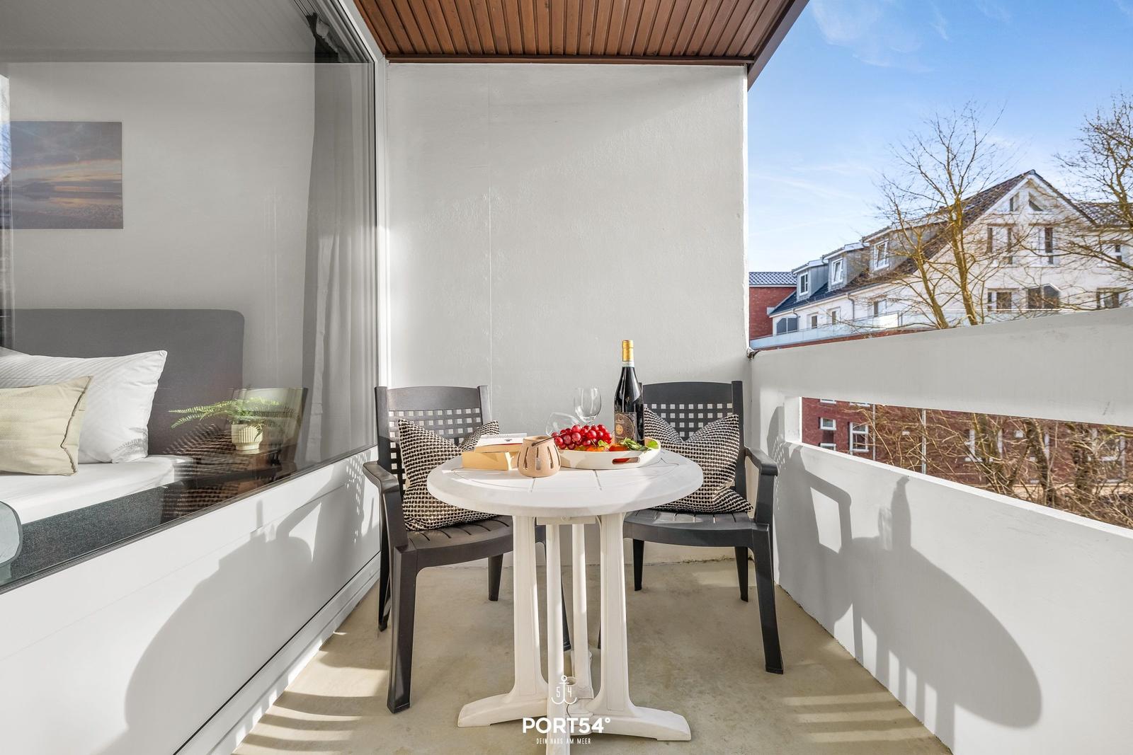 Balcony with table, chairs, and view of buildings. Bed visible through glass.