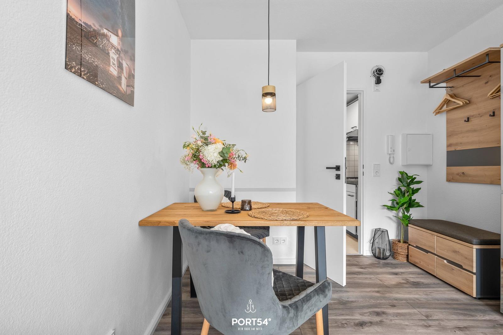Essbereich mit Holztisch, grauer Stuhl und Blumenvase. Hintergrund: Küche und Kleiderschrank.