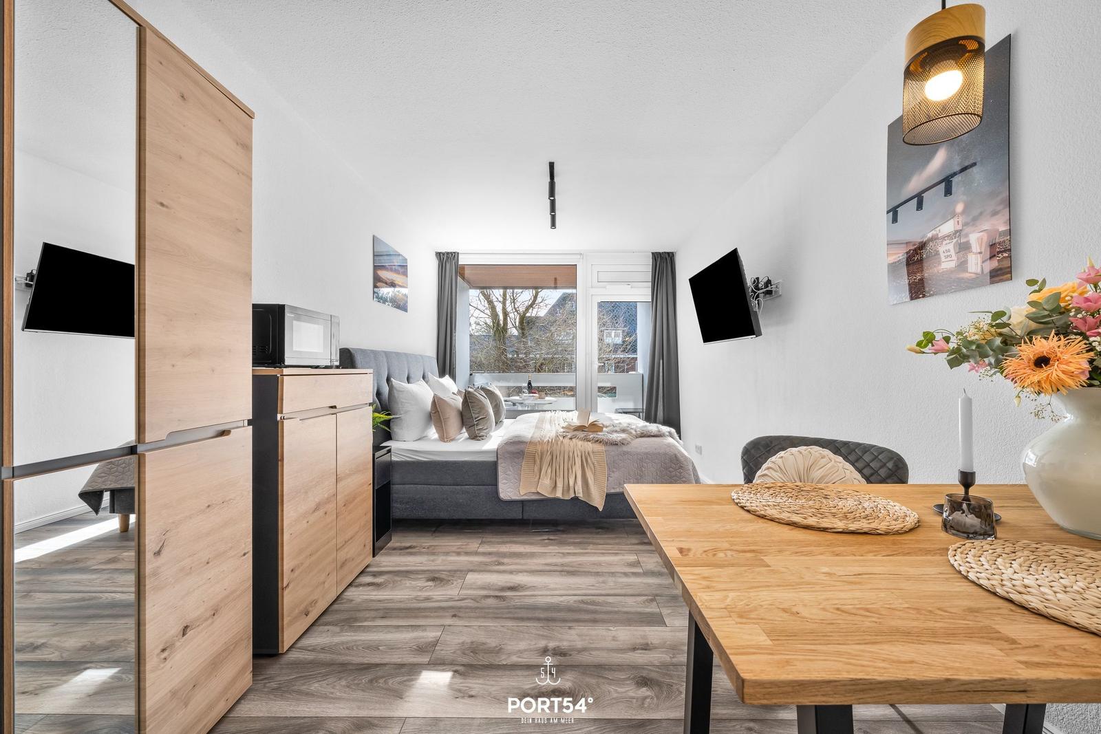 Schlafzimmer mit Bett, TV und Essbereich. Holzfußboden und Fenster zum Balkon.