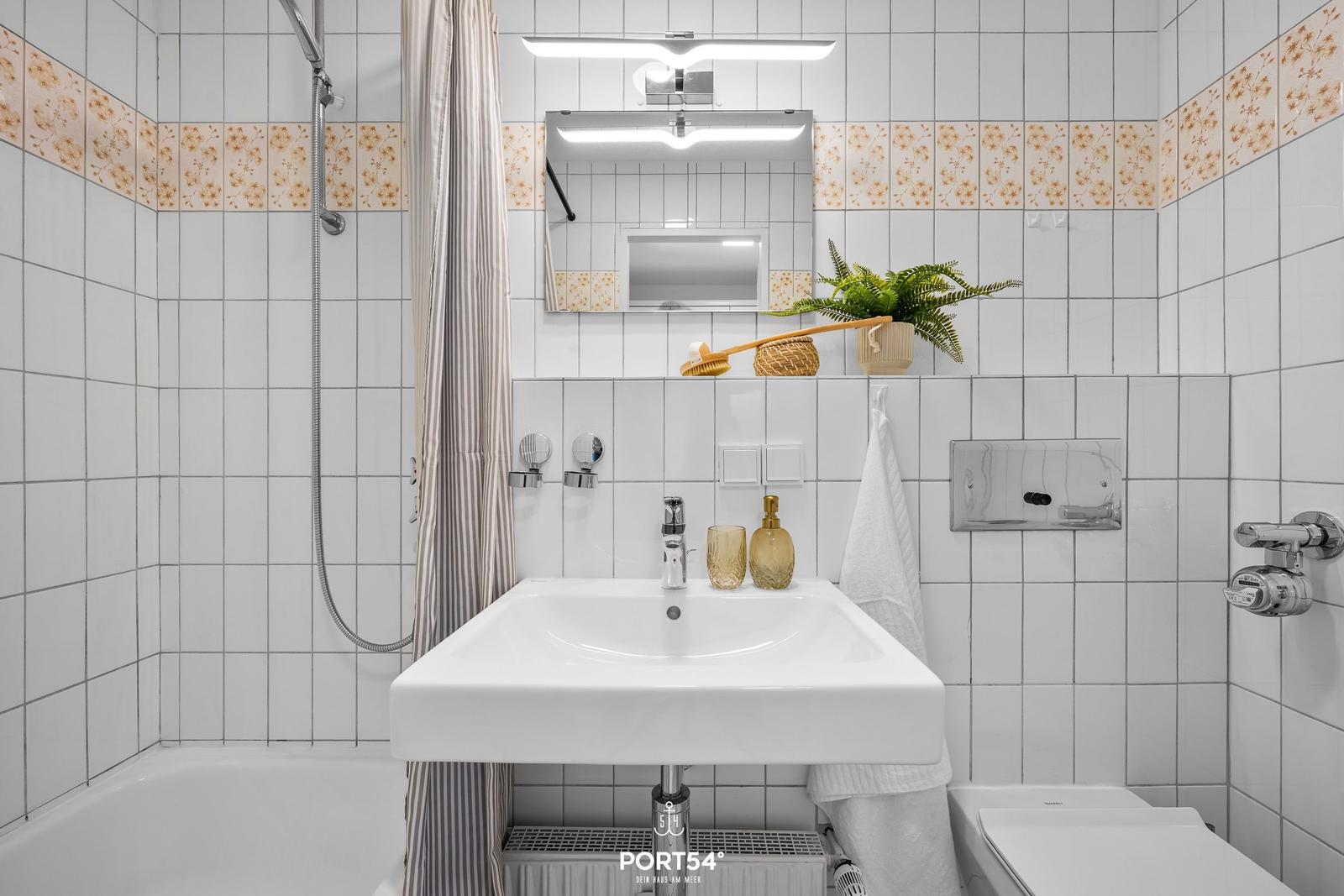 Badezimmer mit Dusche, Waschbecken und Toilette. Weiße Fliesen mit floraler Bordüre.