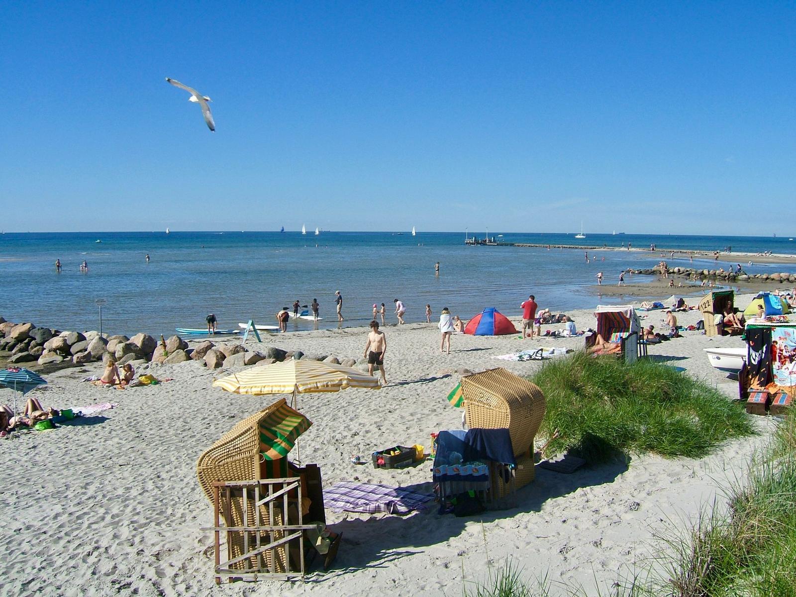 Strand mit Wickerstühlen, Sonnenschirmen und Badegästen am Wasser.