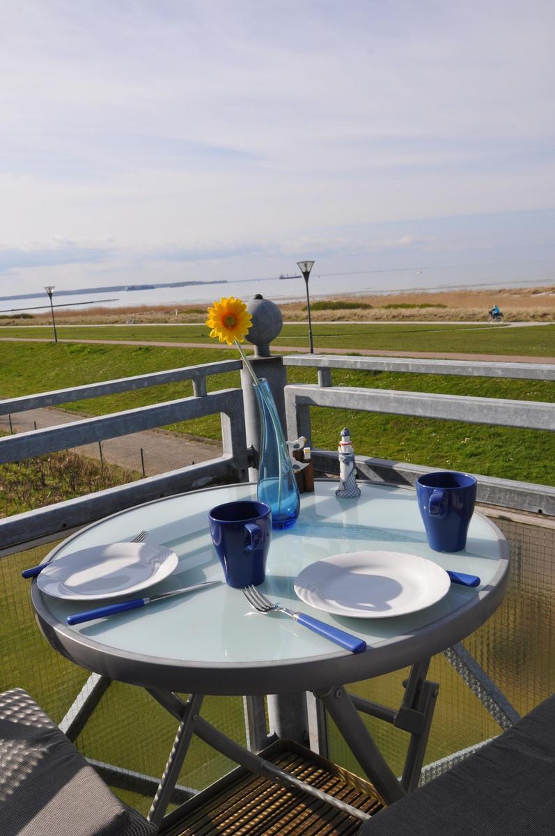 Rundtisch mit Tassen und Blume auf Balkon mit Blick auf Grünfläche und Wasser.