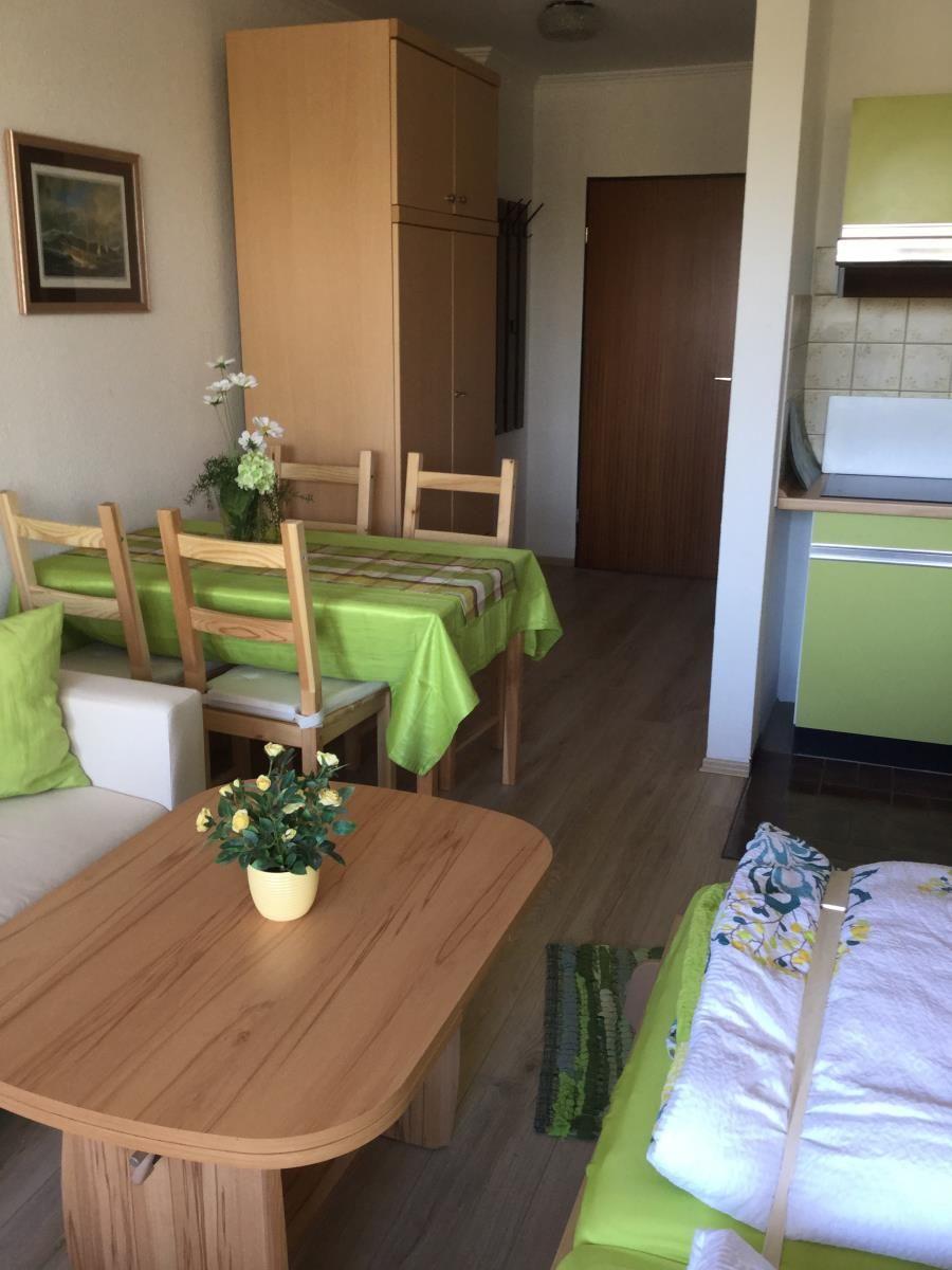 Wohnzimmer mit Esstisch, Sofa und Küchenbereich. Holzmöbel und grüne Tischdecke.