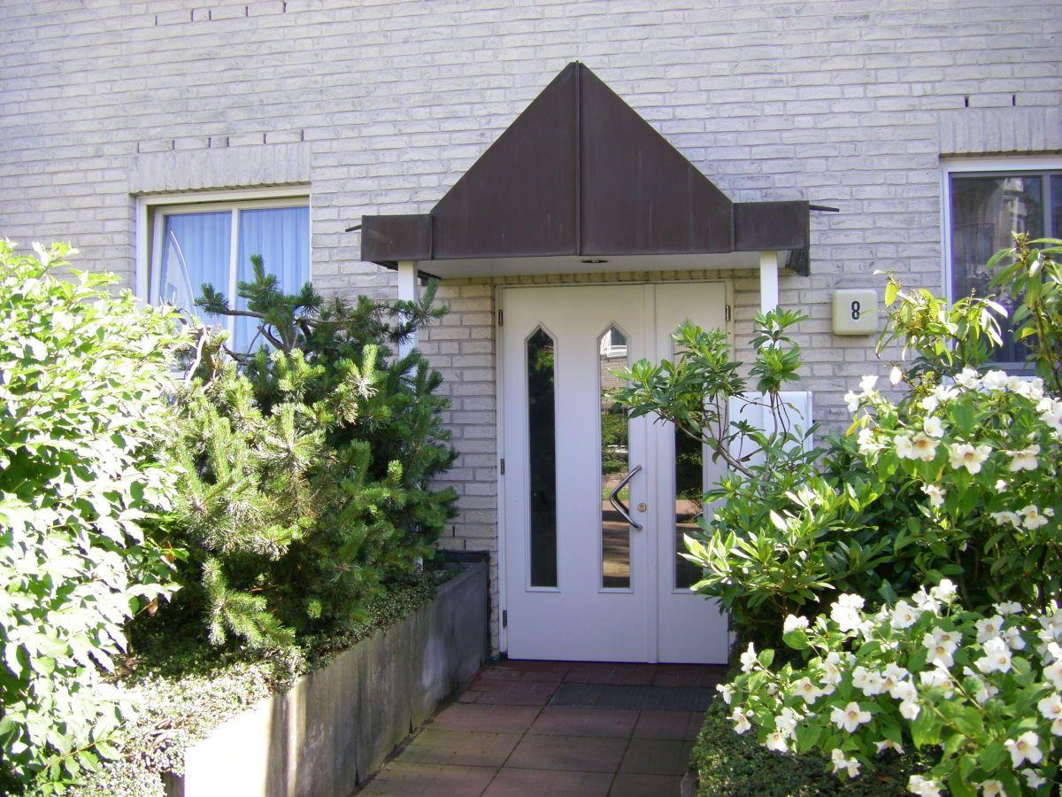 House with white door and dark awning, surrounded by green shrubs and white flowers.