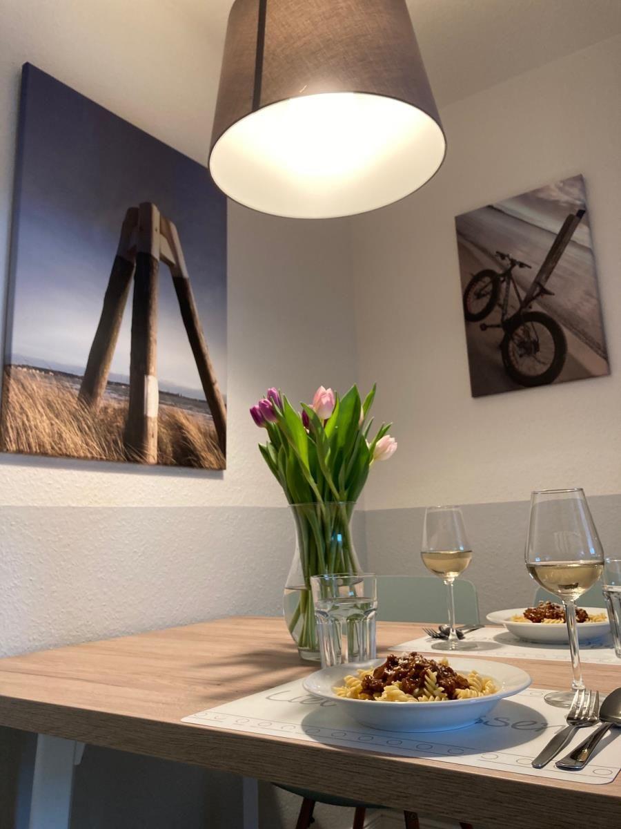 Dining table with pasta, wine, and flowers. Wall art shows beach and bike.