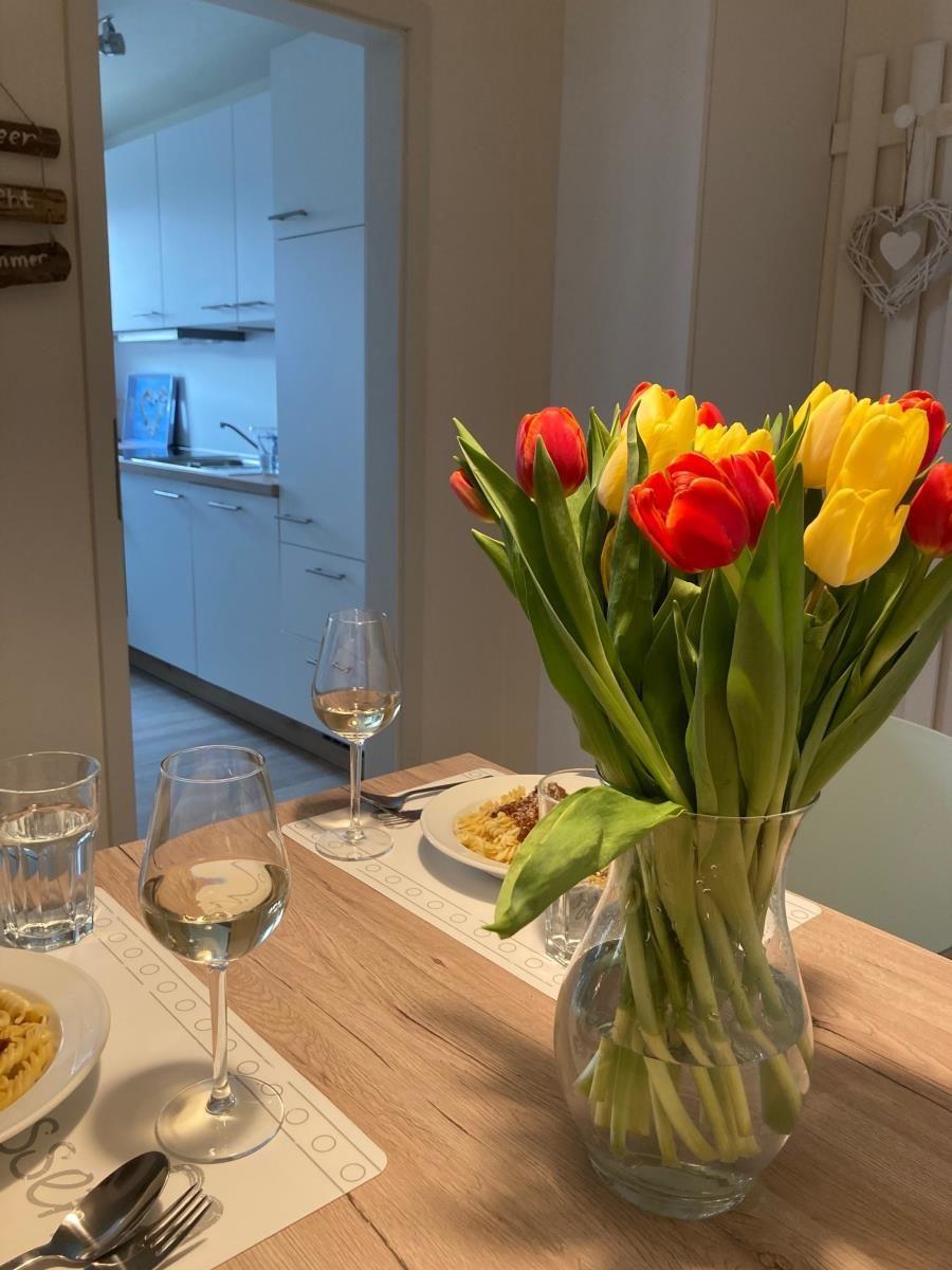 Dining table with flowers, glasses, and food. Kitchen in background.