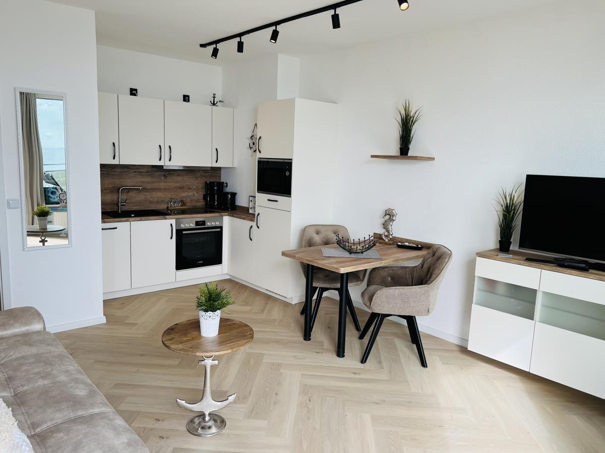 Wohnküche mit Esstisch, TV und Küchenschränken. Holzboden und Spiegel sichtbar.