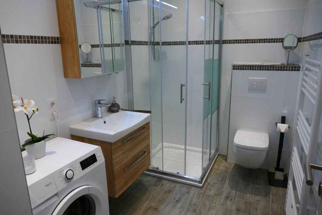 Badezimmer mit Dusche, Waschbecken und Toilette. Waschmaschine und Spiegel sind vorhanden.