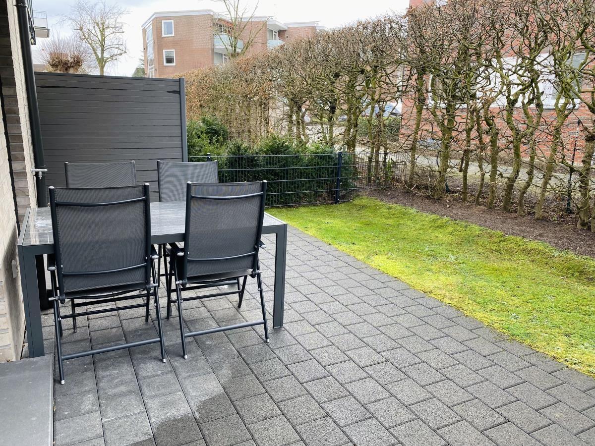 Terrasse mit Tisch und vier Stühlen, gepflastert, mit Rasen und Hecke im Hintergrund.