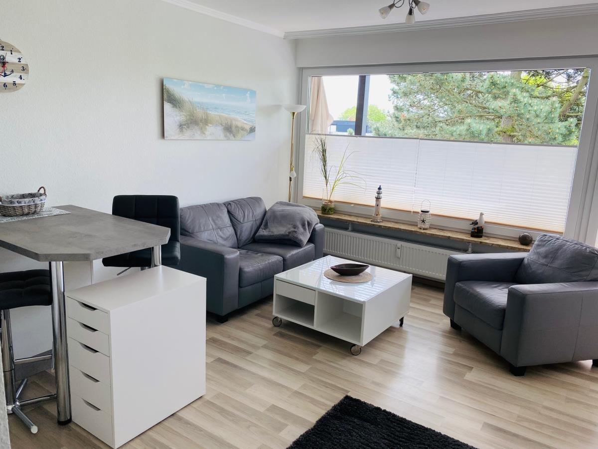 Wohn- und Essbereich mit Sofa, Sessel und Tisch. Große Fenster mit Blick auf Bäume.