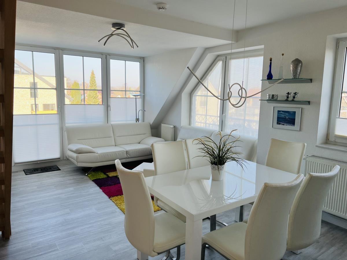 Wohn- und Essbereich mit weißem Sofa, Tisch und Stühlen sowie großen Fenstern.