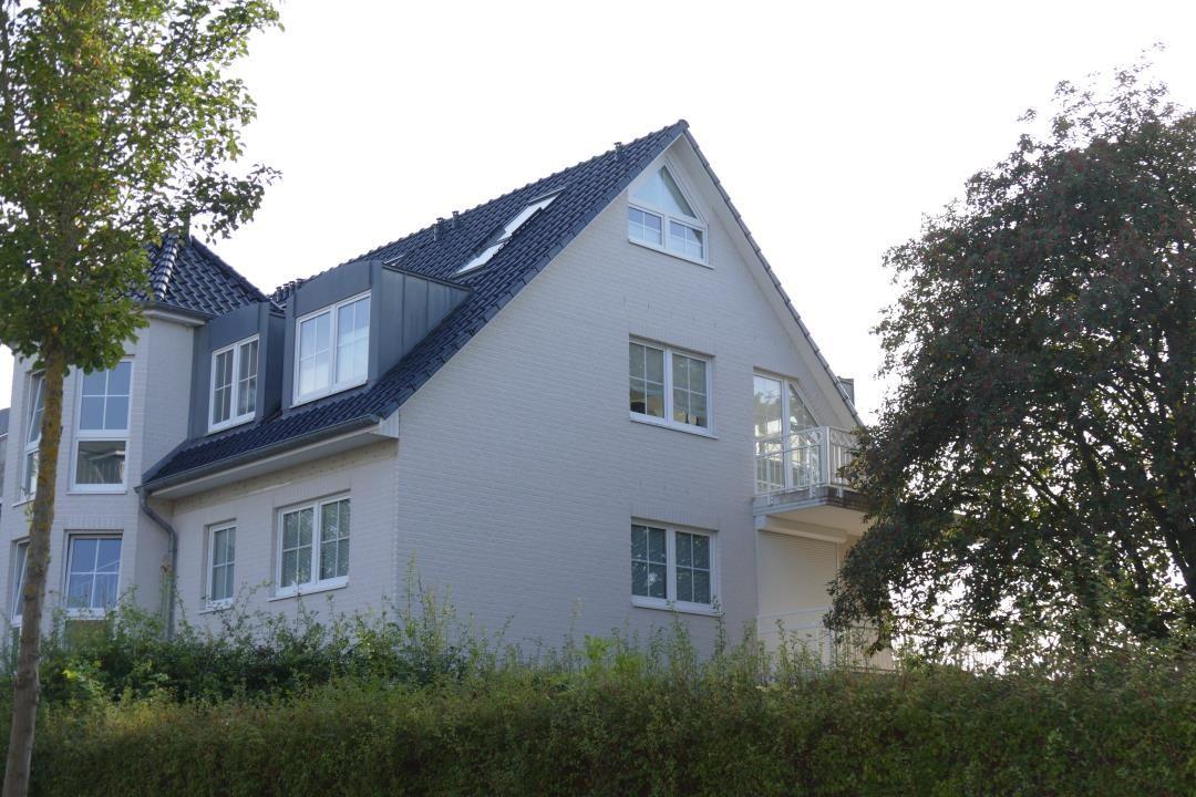 Modernes Haus mit Balkon und Dachfenstern, umgeben von Bäumen und Sträuchern.
