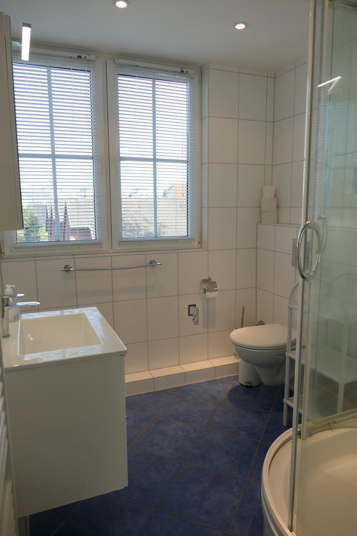 Badezimmer mit Dusche, Toilette und Waschbecken. Blaue Fliesen und Fenster mit Jalousien.