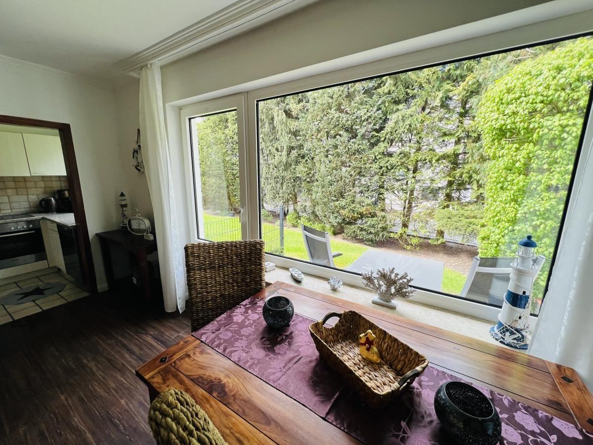 Essbereich mit Tisch, Stühlen und Blick auf Garten. Küchenbereich im Hintergrund.