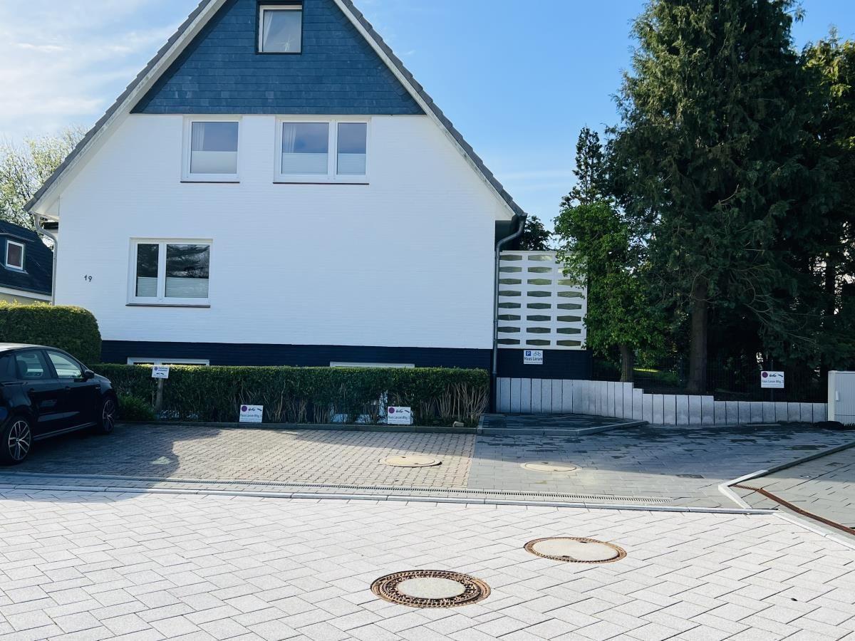 House with white facade and dark roof, paved yard with hedge and tree.