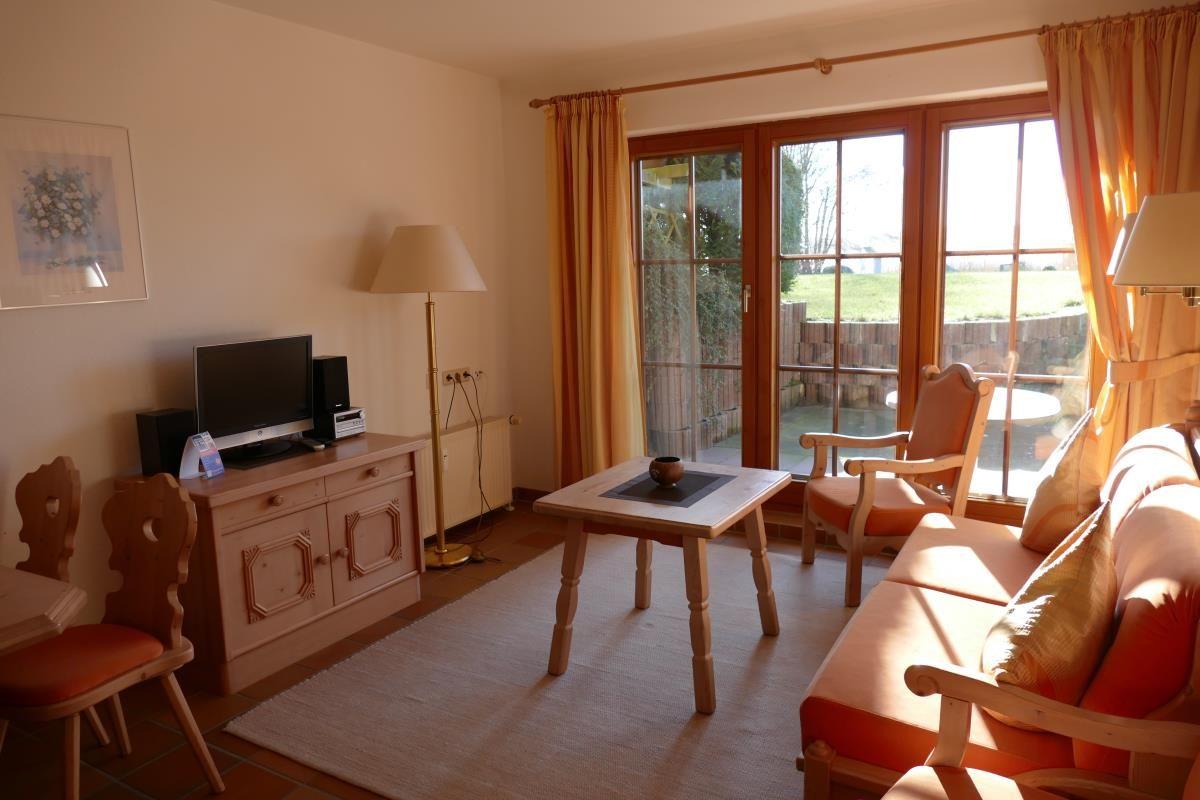 Wohnzimmer mit Sofa, Tisch, Stühlen und TV. Große Glastüren führen auf eine Terrasse.