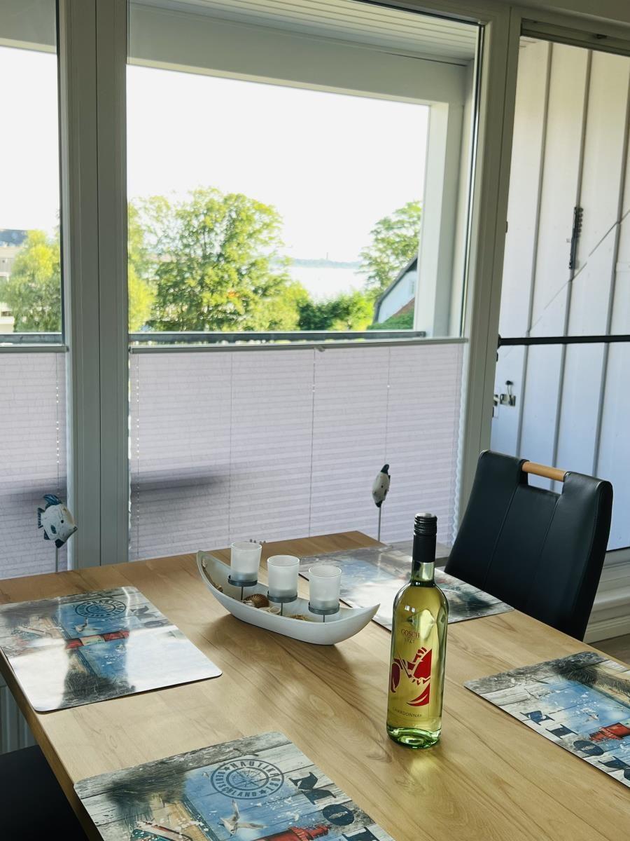Dining table with placemats and wine bottle by window with water view.