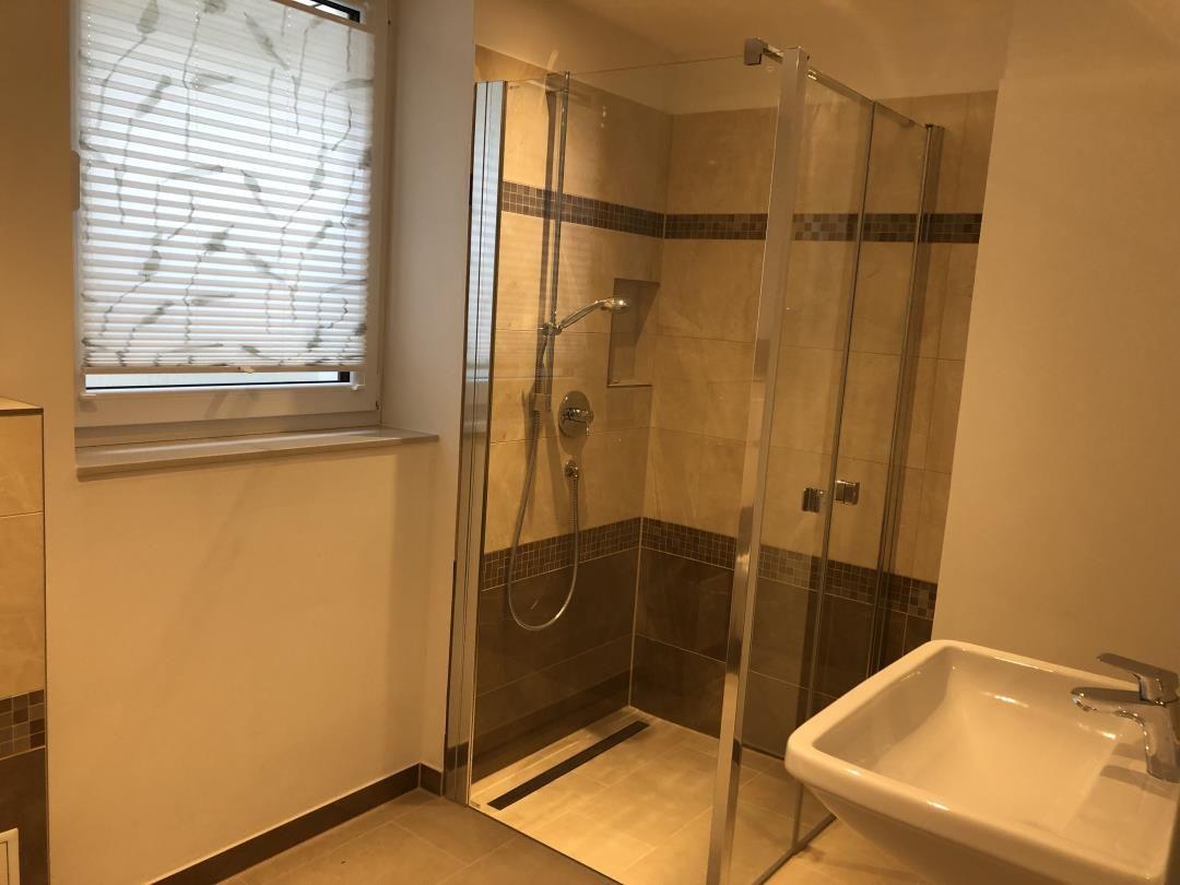 Badezimmer mit Dusche, Waschbecken und Fenster mit Jalousie.