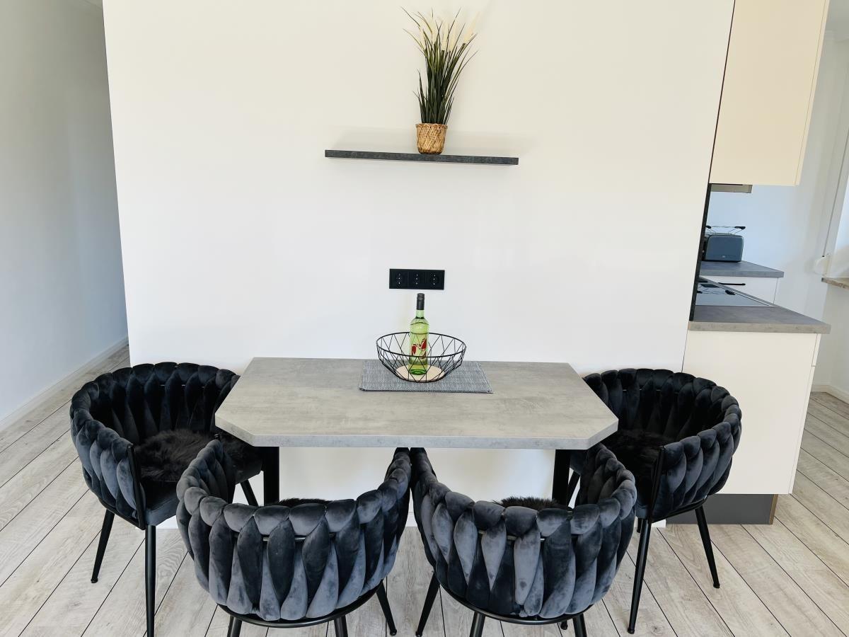 Dining area with table and four chairs, wall shelf with plant, and kitchen area in background.