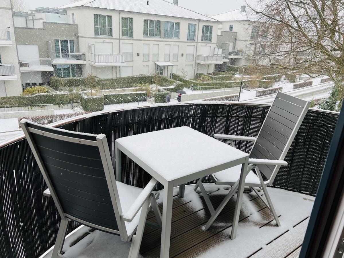 Balkon mit Tisch und zwei Stühlen, bedeckt mit Schnee. Blick auf Wohngebäude und Garten.