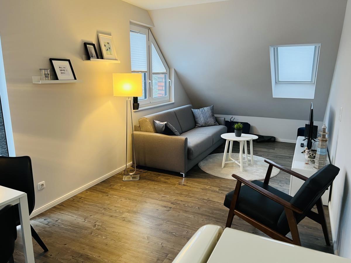 Wohnzimmer mit Sofa, Sessel und Holzboden. Fenster und Stehlampe sichtbar.