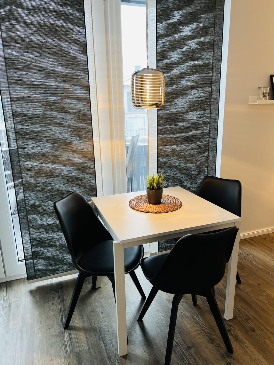 Essbereich mit weißem Tisch, drei schwarzen Stühlen und Pflanze. Hängelampe und Fenster mit Jalousie.