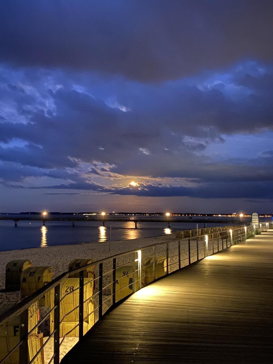 Lit wooden boardwalk along the beach at night with moon and waves.