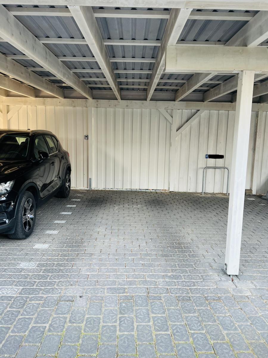Black car parked under a carport with wooden frame and gray paving stones.