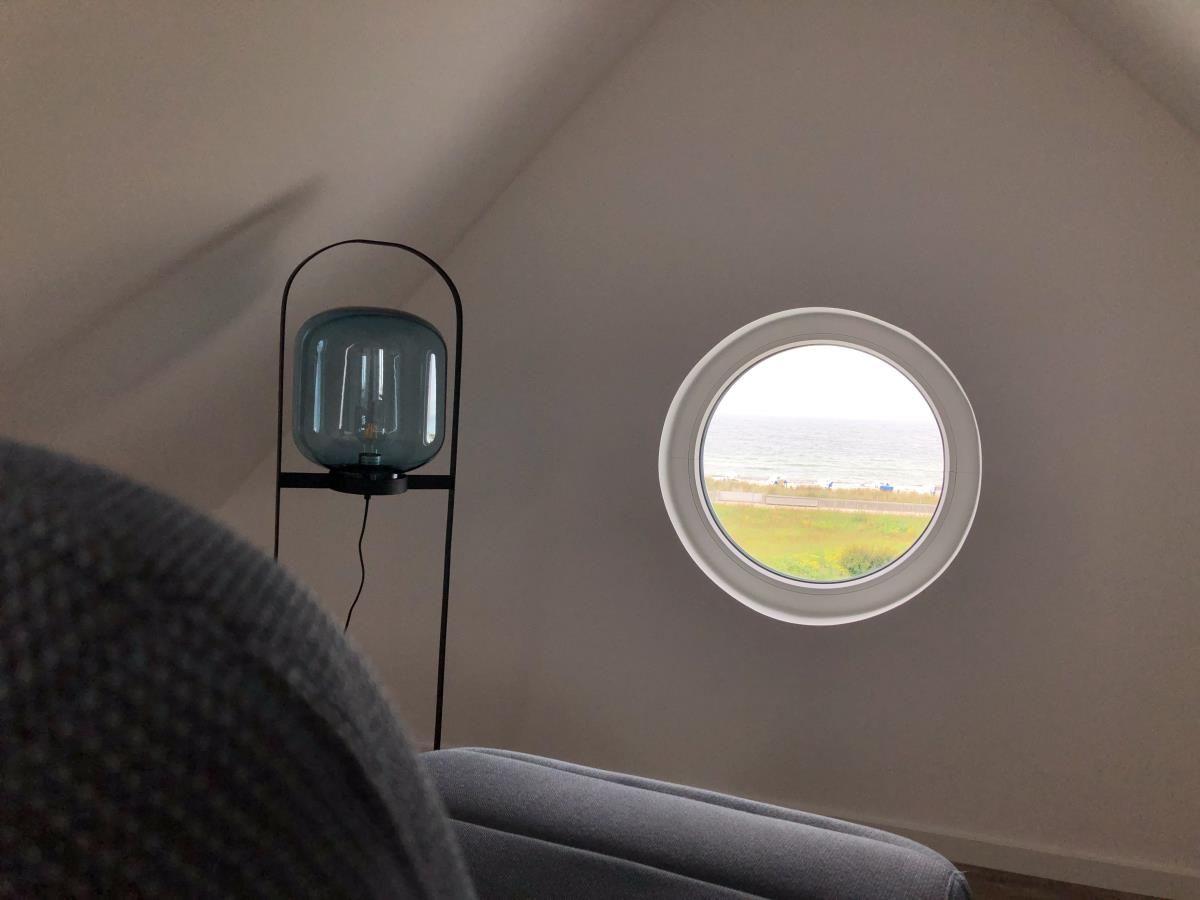 Round window offers view of beach and sea. Next to a gray armchair stands a floor lamp.