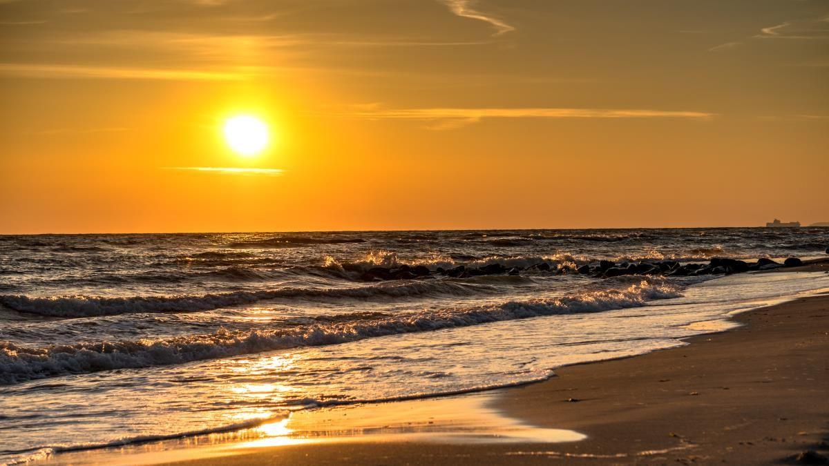 Sonnenuntergang am Strand mit Wellen und einem Schiff im Hintergrund.